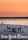 Photo of a romantic table for two beautifully set on a beach at sunset with calm seas and orange skies in the background.